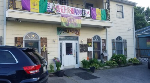 Exterior of a building with a sign reading "Po Boy Factory," decorated with colorful banners and plants.