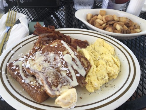 A plate of French toast with syrup, scrambled eggs, bacon, and a side of diced potatoes.