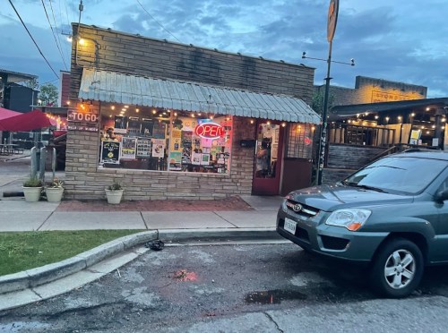 A small, rustic restaurant with an "Open" sign, surrounded by outdoor seating and a parked car nearby.