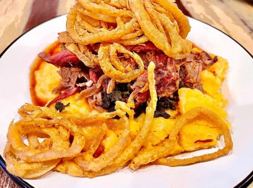 A plate of creamy polenta topped with pulled meat, greens, and crispy onion rings.