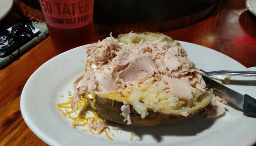 A baked potato topped with shredded chicken and cheese, served on a white plate with a drink in the background.