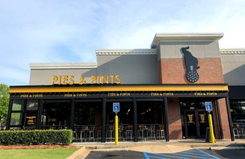 Exterior of a restaurant named "Pies & Pints," featuring a brick facade and large windows, with a dog logo above the entrance.