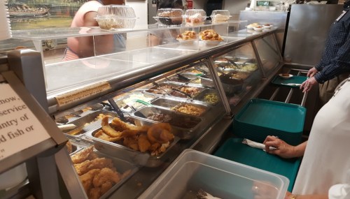 A cafeteria-style food counter with various dishes in metal trays and customers selecting their meals.
