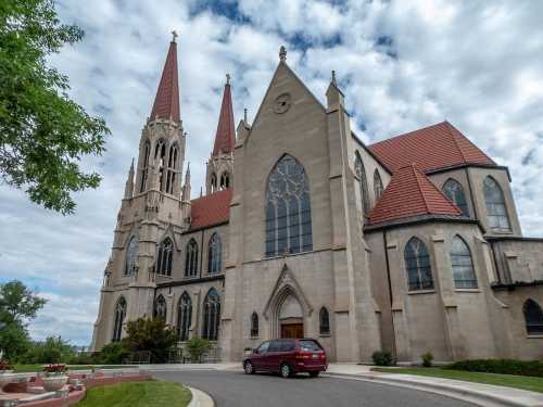 A large stone church with tall spires and stained glass windows, set against a cloudy sky. A car is parked nearby.