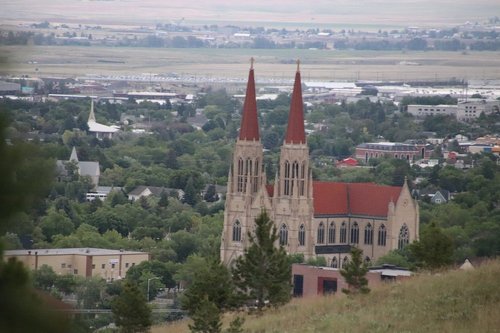 A church with tall spires stands amidst green trees and a cityscape in the background, viewed from a hillside.