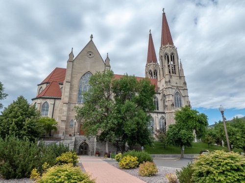 A large stone church with two tall spires, surrounded by greenery and a pathway, under a cloudy sky.