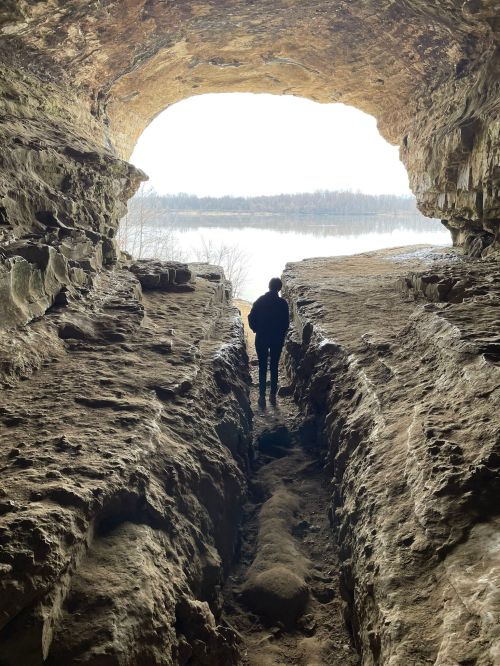 A person stands in a cave, looking out at a river through a large opening, surrounded by rocky walls.