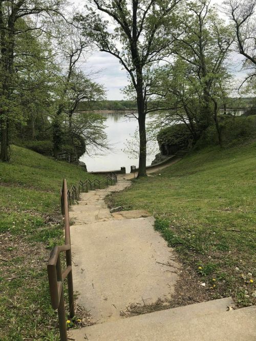 A grassy path leads down to a calm lake, surrounded by trees under a cloudy sky.