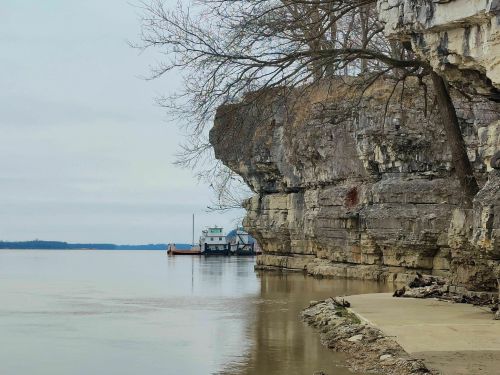 A calm river scene with rocky cliffs, a tree, and a boat docked near the shore under a cloudy sky.