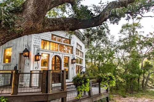 A charming wooden cabin with large windows, surrounded by trees and greenery, featuring a deck with lanterns and plants.