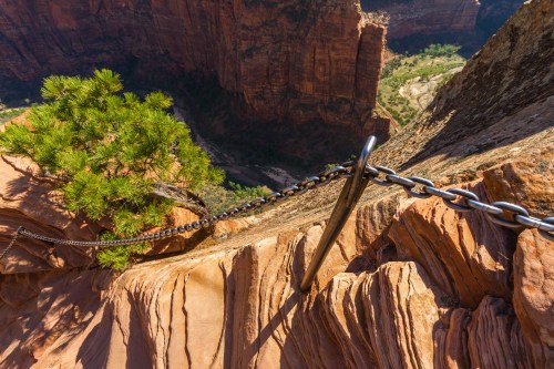 A steep rock face with a metal chain for safety, overlooking a deep canyon and lush valley below.