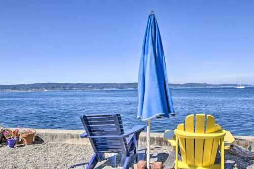Two colorful chairs and a blue umbrella by the water, with a clear sky and distant hills in the background.