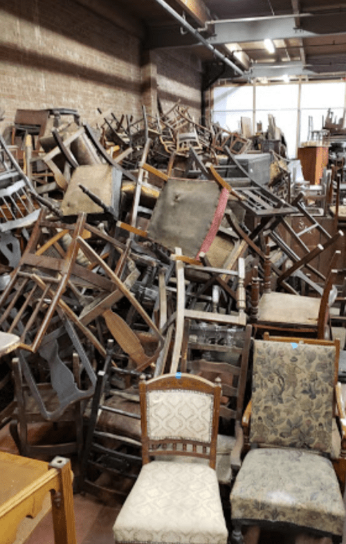 A cluttered room filled with a chaotic pile of wooden chairs and furniture against a brick wall.