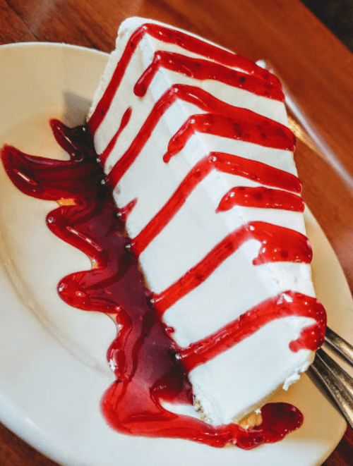 A slice of creamy white cake drizzled with red strawberry sauce on a white plate.