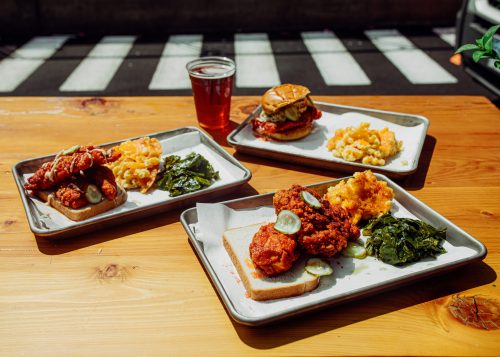 Three trays of Southern-style food featuring fried chicken, mac and cheese, collard greens, and a burger, with a beer.