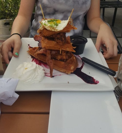 A stack of waffles topped with fried chicken, a fried egg, and a side of sauce, served on a white plate.