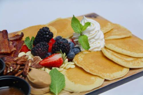 A wooden platter with pancakes, berries, whipped cream, nuts, and bacon, garnished with mint leaves.