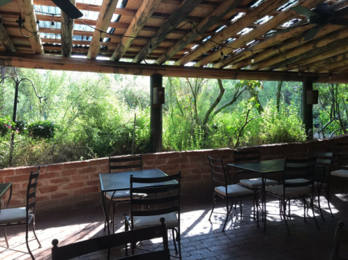 A rustic outdoor dining area with metal tables and chairs, surrounded by lush greenery and wooden beams overhead.