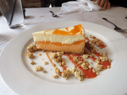 A slice of layered cheesecake with orange topping, drizzled with sauce and surrounded by crumbs on a white plate.