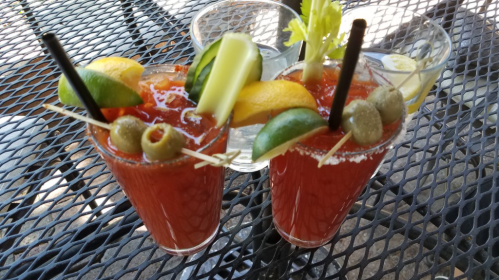 Two colorful cocktails on a table, garnished with olives, lime, cucumber, and orange slices.