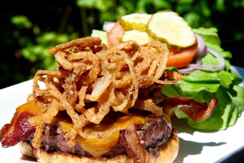 A close-up of a juicy burger topped with bacon, cheese, and crispy onions, served with a side of lettuce, tomato, and pickles.