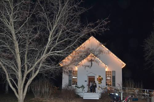 A white house decorated with holiday lights and a wreath, surrounded by bare trees at night.