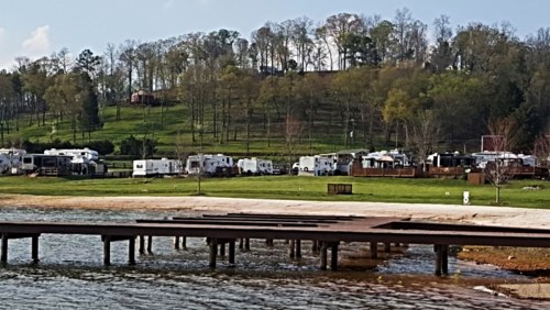 A serene lakeside view with a wooden dock, RVs parked on a grassy hill, and trees in the background under a blue sky.