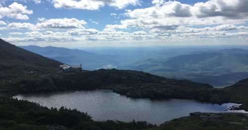 A serene mountain landscape featuring a calm lake, rolling hills, and a cloudy blue sky in the background.