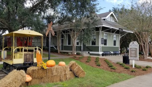 Historic railroad station with a yellow train car, pumpkins, and hay bales in a landscaped area.