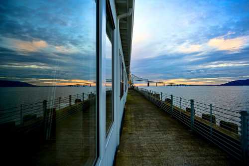 A serene waterfront view at sunset, with reflections on a building and a bridge in the distance.