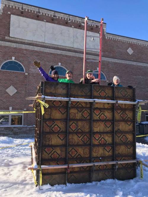 A group of four people wave from atop a large wooden structure in a snowy outdoor setting.