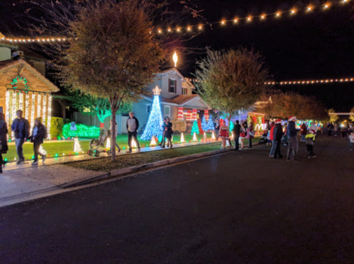 A festive street scene with houses decorated in colorful holiday lights and people walking along the sidewalk.