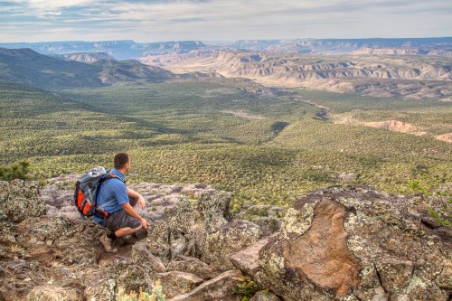 A hiker sits on a rocky ledge, overlooking a vast green valley and distant mountains under a cloudy sky.