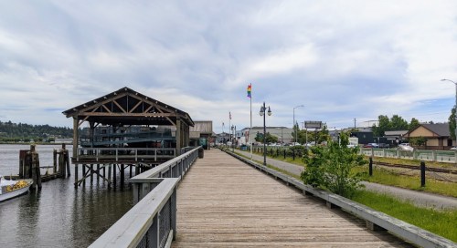A wooden boardwalk along a waterfront with a dock, buildings, and flags, under a cloudy sky.