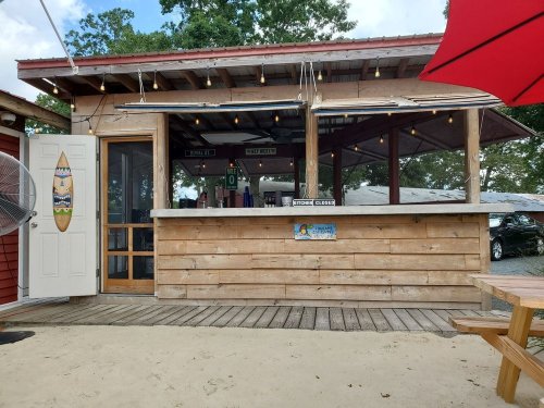 A wooden outdoor bar with a closed kitchen sign, surrounded by trees and a sandy area, under a partly cloudy sky.