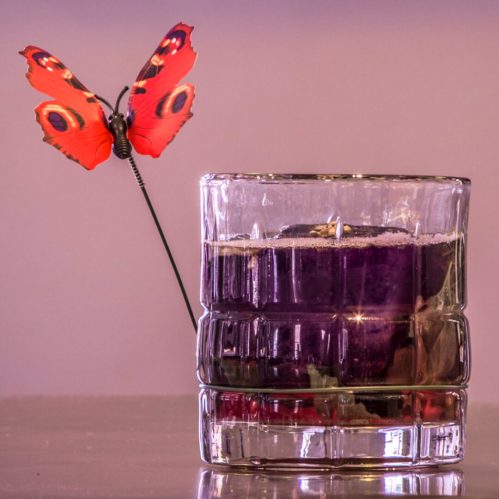 A vibrant red butterfly perched on the rim of a glass candle holder filled with purple wax.