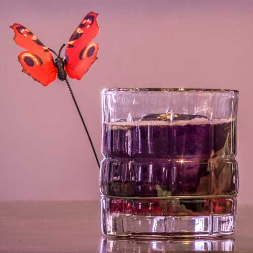 A vibrant red butterfly perched on the rim of a glass candle holder filled with purple wax.