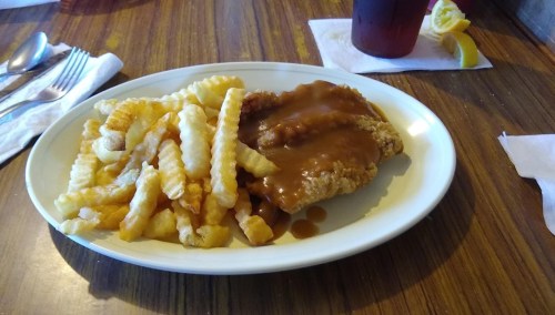 A plate of crispy fried chicken smothered in gravy, served with crinkle-cut fries and a drink on the side.