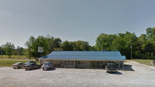 A single-story building with a blue metal roof, surrounded by trees and parked cars in a gravel lot.