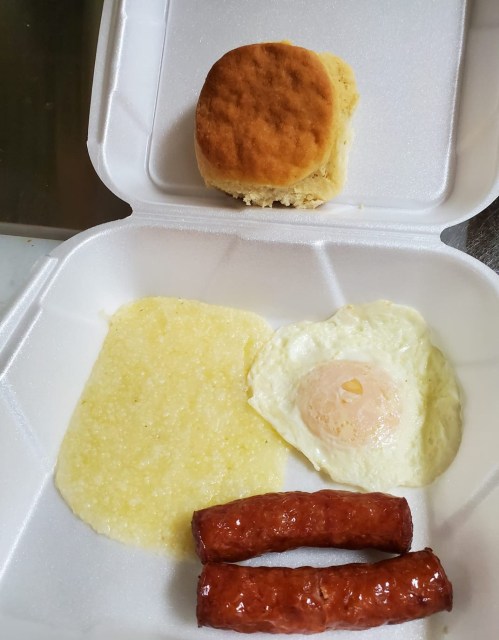 A takeout container with a biscuit, grits, a fried egg, and two sausage links.