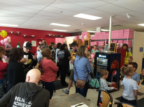 A busy bakery filled with people, balloons, and pink boxes, creating a lively atmosphere.
