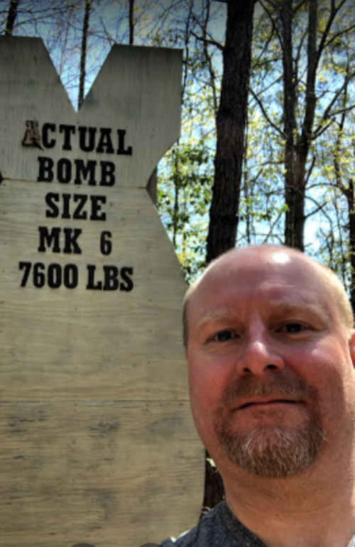 A man takes a selfie in front of a large sign that reads "Actual Bomb Size MK 6 7600 lbs" in a wooded area.