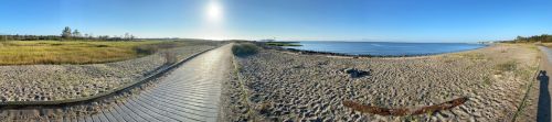 A panoramic view of a sandy beach with a wooden walkway, calm water, and clear blue skies.
