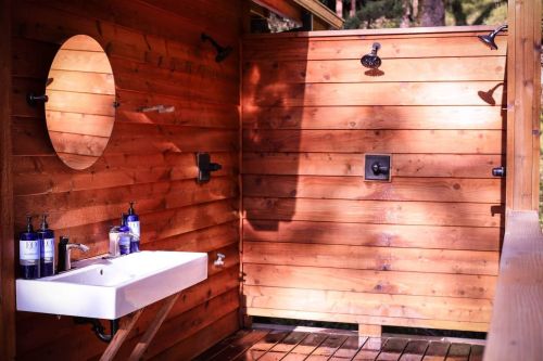 A rustic outdoor shower area with wooden walls, a round mirror, and a sink, surrounded by trees and natural light.