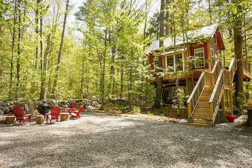 A cozy red treehouse surrounded by lush green trees, with a gravel area and red chairs for outdoor seating.