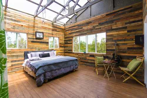 Cozy bedroom with wooden walls, large windows, a bed, and a small table with chairs and a guitar.