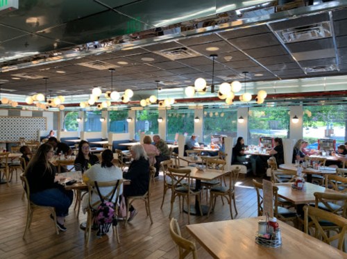 A bright restaurant interior with wooden tables, chairs, and groups of people dining and chatting.