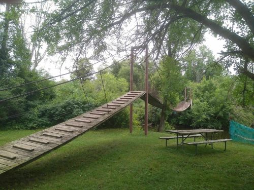 A wooden suspension bridge connects two sides of a lush, green park, with a picnic table in the foreground.