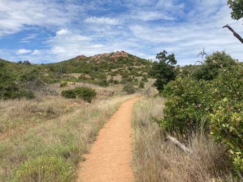A dirt path winds through grassy terrain, leading towards a rocky hill under a partly cloudy sky.