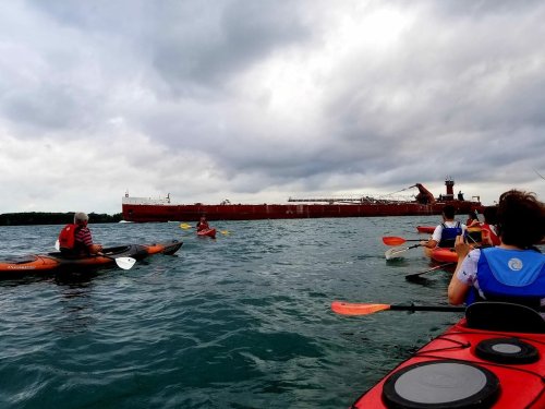 A group of kayakers paddles in the water near a large cargo ship under a cloudy sky.
